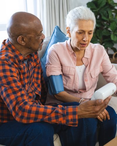 Diverse senior man testing blood pressure of senior woman in sunny living room. Lifestyle, retirement, senior lifestyle, togetherness and domestic life, unaltered.